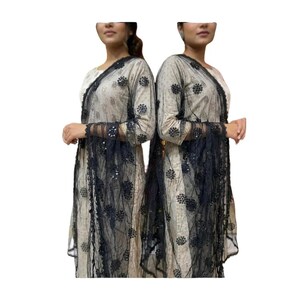 May include: Two women wearing matching beige and black outfits. The outfits include a long-sleeved top and a sheer black shawl with floral patterns and sequins. The outfits have a traditional design.