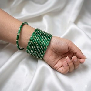 May include: A collection of green glass bangles with gold-colored accents. The bangles are stacked on a wrist, with a single bangle on the forearm. The jewelry is displayed on a white, silky fabric.
