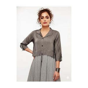 May include: A gray collared shirt with three-quarter sleeves and a button-down front. The shirt is paired with a gray and white patterned dress. A silver cuff bracelet is visible on the wrist.