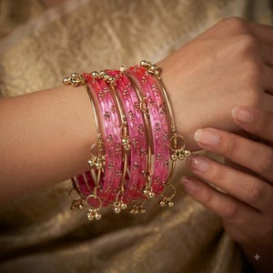 May include: A set of translucent hot pink bangles featuring a textured surface with embedded gold-toned glitter accents. Each bangle is adorned with small, dangling gold-toned metal charms that hang from thin circular loops attached to the bands.