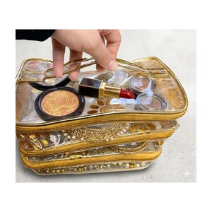 May include: A clear, gold-trimmed makeup bag with multiple compartments, holding various cosmetics. The bag contains a compact with a gold design, a red lipstick, and eyeshadow. The bag is held by a hand.