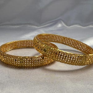 May include: Two gold-colored bangles with intricate openwork designs. The bangles are round and have a textured surface with small, evenly spaced holes. They are displayed on a white, slightly wrinkled fabric.