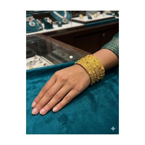 May include: A set of gold bangles adorns a wrist, showcasing intricate patterns and small red gemstones. The bangles are stacked, creating a bold statement. The jewelry is displayed on a teal surface, with other jewelry visible in the background.