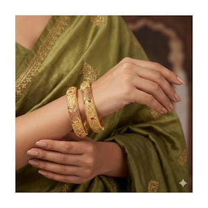 May include: Two gold bangles with intricate floral and geometric designs, adorned with small red gemstones. The bangles are worn on a wrist, set against a backdrop of a green fabric with gold embroidery.