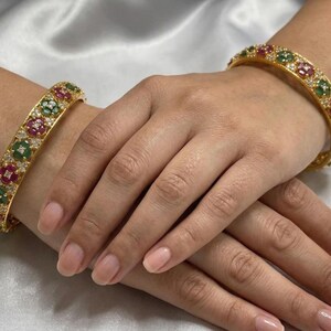 May include: Two gold bangles, each adorned with alternating red, green, and clear gemstones. The bangles are displayed on a white satin surface. The gemstones are arranged in a floral pattern.