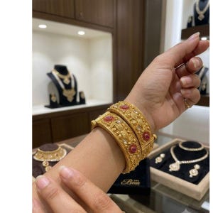 May include: Two gold bangles featuring intricate embossed floral patterns, small clear stones, and circular red accents. The bangles have a hinged design with a visible clasp mechanism, showcasing a textured finish and a wide, ornate band structure.