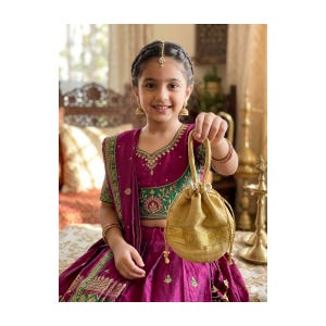 May include: A young person wearing a traditional Indian outfit, holding a gold drawstring bag. The outfit is a vibrant magenta with green and gold embroidery. The bag has a gold band around the bottom.