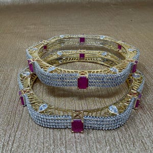May include: Two gold-toned bangles with a geometric design. The bangles are encrusted with small, clear stones and feature rectangular ruby-colored accents. The jewelry is displayed on a gold-colored surface.