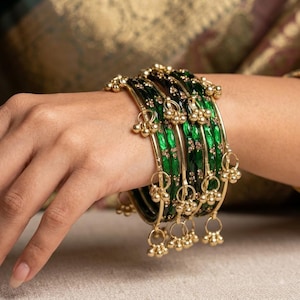 May include: A set of stacked bangles featuring a vibrant emerald green enamel finish with gold-toned metal accents. Each bangle is adorned with small, dangling gold-toned charms and embedded with tiny, sparkling clear stones in a floral pattern.