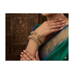 May include: Two gold-toned bangles with a woven design and red accents are displayed on a person's arm. The bangles are worn with a green and blue garment and gold jewelry, including a necklace and rings. The background is a dark, patterned surface.