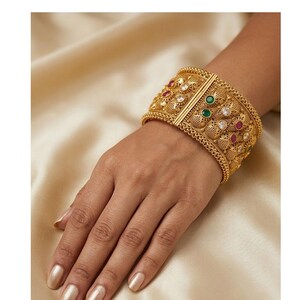 May include: A wide gold cuff bracelet featuring intricate filigree metalwork, a central vertical bar, and embedded round-cut gemstones in red, green, and clear white. The textured surface displays a repeating pattern of small metallic beads.