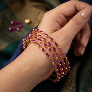 May include: Gold-toned bangles with marquise-cut ruby-colored stones. The bangles are displayed on a hand, showcasing the intricate design and vibrant gemstones. The jewelry is set against a backdrop of blue and green fabric.