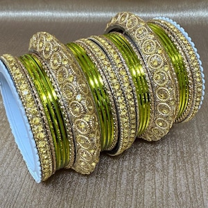 May include: A stack of gold-colored bangles with intricate patterns and embedded yellow gemstones. The bangles have a bright green inner layer, and the edges are lined with white. The jewelry is displayed on a textured, golden-brown surface.