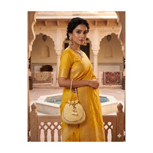May include: A woman wearing a yellow sari with gold jewelry, including earrings and a necklace. She is holding a small, round, cream-colored purse with a pearl handle and tassel details. The background features an ornate architectural setting.