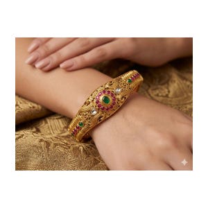 May include: Gold-toned bangle bracelet with intricate filigree design. The bracelet features a central design with a green gemstone surrounded by red and white gemstones. The bracelet is worn on a wrist.