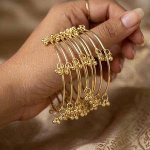 May include: A hand displays a collection of gold bangles. Each bangle is thin and adorned with small, spherical embellishments. The bangles are a warm gold color, reflecting light. The background is a soft, neutral tone, highlighting the jewelry.
