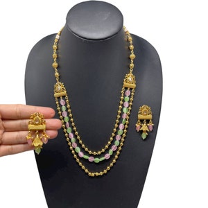 May include: A gold necklace with three strands of beads. The beads are alternating pink and green. The necklace has a gold pendant with a small white stone. The necklace comes with a pair of matching earrings with pink and green beads.