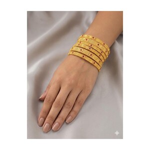 May include: A set of six gold bangles with intricate patterns and small red accents. The bangles are stacked on a wrist, showcasing their design and color against a neutral background. The jewelry is likely for special occasions.