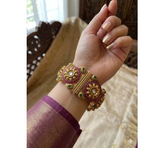 May include: A wide gold-tone cuff bracelet featuring two prominent circular floral motifs. The design is accented with deep pink stones and small green stone accents, with a central white stone in each flower. The metal has a textured, matte finish.