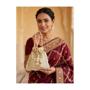 May include: A woman holding a light green drawstring bag with floral embroidery and a pearl handle. The bag has a cream-colored lace trim and gold tassels. She is wearing a maroon sari with gold embroidery and a pearl necklace and earrings.