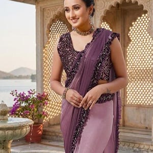 May include: A woman wearing a mauve saree with a sequined top and ruffled edges. The saree has a draped design and is paired with matching jewelry. The background features an ornate architectural structure and a water feature.