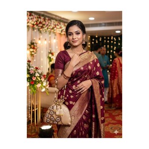 May include: A woman wearing a maroon sari with gold detailing and a matching blouse. The sari features a patterned design. She is holding a beige drawstring bag with gold and pearl embellishments. The background includes floral decorations and soft lighting.