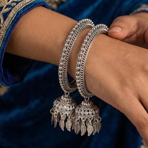 May include: Two silver bangles with intricate beaded and filigree designs. Each bangle features a bell-shaped charm with dangling leaf accents. The jewelry is displayed against a blue background.