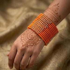 May include: A stack of orange bangles with rhinestone accents. The bangles are wrapped in orange thread and have a gold-colored trim. The image shows a hand with henna art.