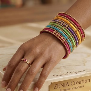 May include: A stack of colorful bangles in shades of pink, yellow, green, blue, and purple, adorned with gold-colored beads. A gold ring with a pink gemstone is on the ring finger. The bangles are on a person's wrist.