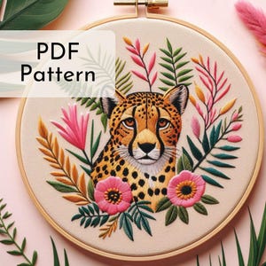 May include: Embroidered hoop art featuring a cheetah surrounded by tropical foliage and pink flowers. The hoop is made of wood and has a beige fabric background. The text "PDF Pattern" is displayed in the top left corner of the image.