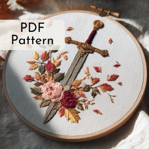 Può includere: Ricamo su telaio raffigurante una spada con elsa dorata e impugnatura bordeaux, circondata da ricami floreali nei toni del rosa, rosso e oro. Il testo "PDF Pattern" è visualizzato in alto a sinistra. Il ricamo è realizzato su un telaio di legno.