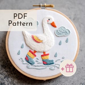 Könnte beinhalten: Gesticktes Bild in einem Holzrahmen, das eine weiße Gans mit bunten Gummistiefeln zeigt, die im Regen spazieren geht. Das Design umfasst eine Wolke und Regentropfen, mit dem Text "PDF Pattern".