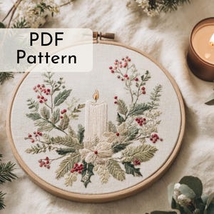 Puede incluir: Un patrón PDF para un bordado en bastidor. El diseño presenta una vela central rodeada de vegetación y bayas rojas. El bordado está en un bastidor de madera, creando un aspecto festivo y artesanal. El texto "PDF Pattern" está superpuesto en la imagen.