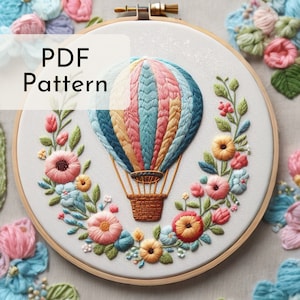 Puede incluir: Un globo aerostático colorido con una cesta, rodeado de una corona de flores bordadas en tonos de rosa, azul y verde. El bordado se muestra en un aro de bordado de madera. El texto "PDF Pattern" se muestra en la esquina superior izquierda de la imagen.