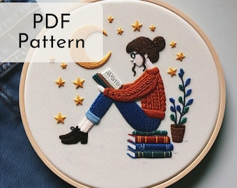 May include: Embroidered hoop art featuring a woman reading a book under a crescent moon with stars. She is sitting on a stack of books with a potted plant beside her. The embroidery is done in shades of blue, red, brown, and yellow.