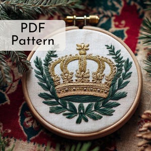Op de afbeelding: Geborduurd PDF-patroon in een houten ring. Het ontwerp toont een gouden kroon omringd door groene bladeren. De tekst "PDF Pattern" staat in een wit kader. De borduursels zijn op een crèmekleurige stof.