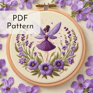 Op de afbeelding: Een PDF-patroon voor borduurwerk met een paarse fee met vleugels, die in een cirkel van paarse bloemen en groene bladeren staat. Het ontwerp is in een houten borduurring.