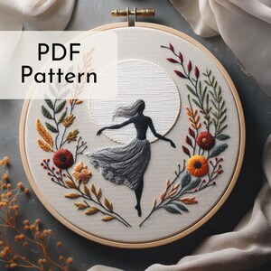 May include: Embroidered hoop art featuring a silhouette of a woman dancing in front of a full moon, surrounded by a wreath of colorful flowers and leaves.