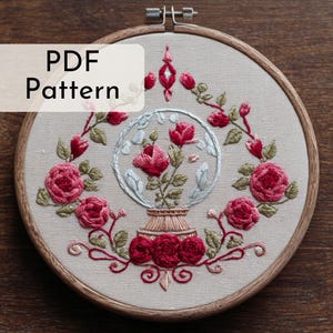 May include: Embroidered floral design in a wooden hoop. The design features red roses, green leaves, and a glass globe with flowers. The text "PDF Pattern" is in the upper left corner.