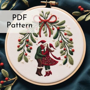 May include: Embroidered hoop art featuring a couple standing under a mistletoe sprig with red berries. The couple is wearing red and green clothing and Santa hats. The background is white with a subtle sparkle effect. The text "PDF Pattern" is in the top left corner.