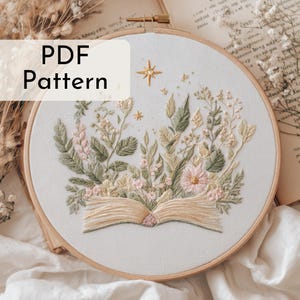 Könnte beinhalten: Gesticktes Blumendesign in einem Holzreifen. Die Stickerei zeigt ein offenes Buch mit Blumen und Blättern in Grün-, Rosa- und Cremetönen. Der Text "PDF Pattern" befindet sich in einem weißen Feld oben links.