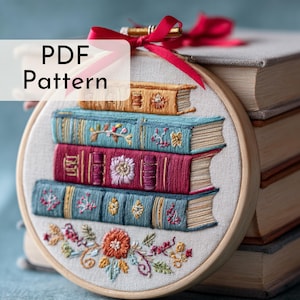 May include: An embroidered hoop art piece featuring a stack of colorful books. The books are in shades of teal, burgundy, and gold, with detailed floral embroidery. The text "PDF Pattern" is displayed on the upper left. A red ribbon is tied at the top.