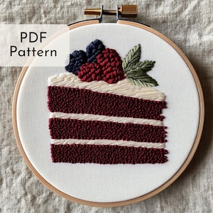 May include: A red velvet cake slice with white frosting and a berry topping. The cake is embroidered on a white fabric stretched over a wooden embroidery hoop.