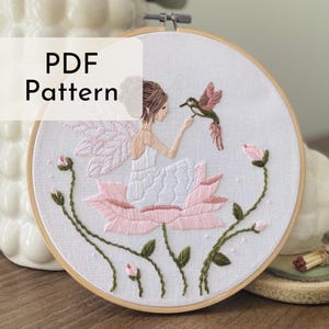 May include: A PDF pattern for embroidery featuring a fairy sitting on a pink flower with a hummingbird. The fairy has pink wings and is wearing a white dress. The hummingbird is pink and green.