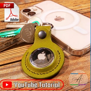 Op de afbeelding: Een groene leren AirTag-sleutelhanger met een gestikte rand en een zilveren Apple-logo. De sleutelhanger ligt naast een transparante telefoonhoes met een witte ring aan de achterkant. De afbeelding bevat de tekst "YouTube Tutorial" en "LEATHER PATTERN".