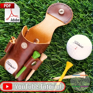 May include: A brown leather golf tee holder with a Titleist golf ball, green and yellow tees. The holder has a snap closure and a loop for attaching to a golf bag. The image also includes text that says "The Golfer's Leather Essential" and "Leather Pattern".
