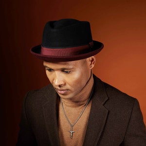 Op de afbeelding: Een man draagt een zwarte fedora met een donkerrode band en een bruine turtleneck trui. Hij draagt een zilveren ketting met een kruis hanger.