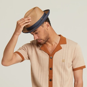 May include: A fedora hat with a tan woven crown and a navy blue brim. The hat has a dark brown band and a button detail. The person in the image is wearing a tan short-sleeved shirt with brown trim and buttons.