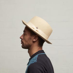 May include: A cream-coloured fedora hat with a flat top and a ribbon band. The hat has a small, decorative bow and a silver-coloured button. The brim is slightly upturned. The hat is made of a felt-like material.