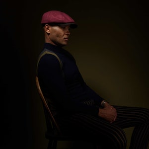 May include: A man wearing a burgundy newsboy cap sits in a chair, wearing a dark blue sweater with a yellow and white stripe pattern and striped pants.